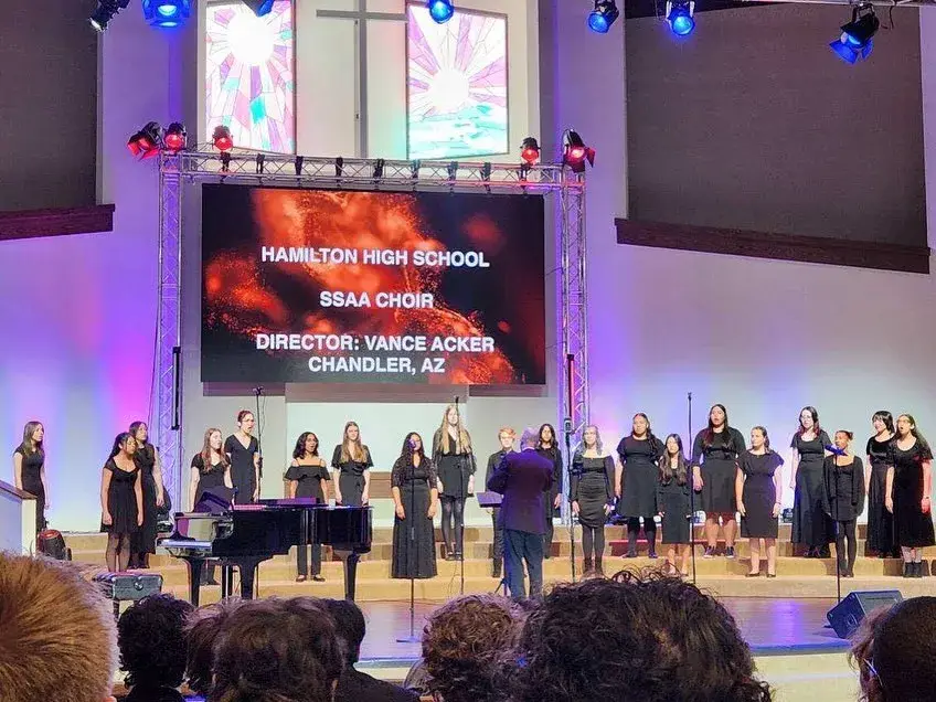 Eurydice in black dresses performing on stage in Anaheim under a screen reading Hamilton High School SSAA Choir